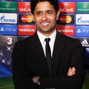 Uśmiechnnięty Nasser Al-Khelaifi w czarnym garniturze na tle ścianki reklamowej prezentującej logo piłkarskiej Ligi Mistrzów oraz jej sponsorów