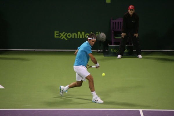 Na zdjęciu Roger Federer na korcie o twardej nawierzchni, w niebieskiej koszulce polo i białych szortach, szykuje się do uderzenia piłki z bekhendu