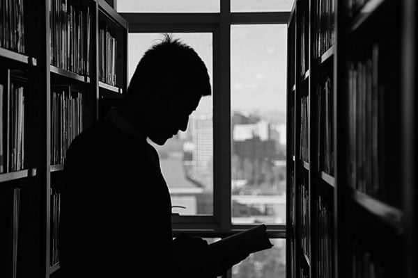 Czarno-białe zdjęcie mężczyzny ukazanego z profilu od pasa w górę, stojącego między dwoma bibliotecznymi regałami i trzymającego w rękach otwartą książkę. Widoczny jest jedynie zarys sylwetki postaci na tle jasnego okna